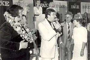 Johnny Reimar taler i mikrofon ved en fest eller reception i sort-hvid. Han er klædt i lys jakke og holder mikrofonen op mod en blomsterkransprydet gæst til venstre. I baggrunden ses Faxe-logoer og flere tilskuere. Billedet er fra 1980'erne.